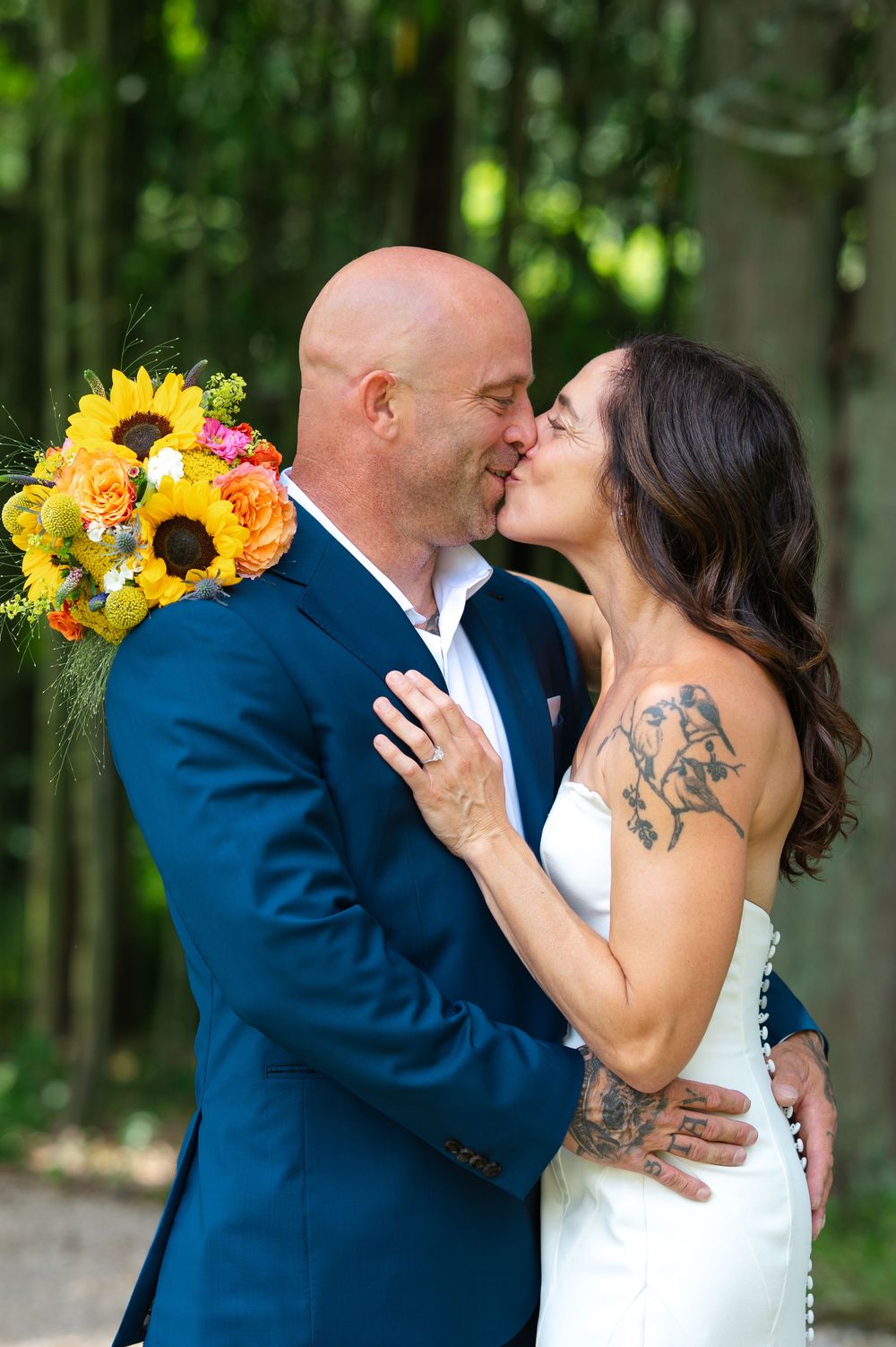 Bride and groom sharing a kiss outdoors, holding a vibrant sunflower bouquet, bride in white dress with tattoos, groom in navy suit.