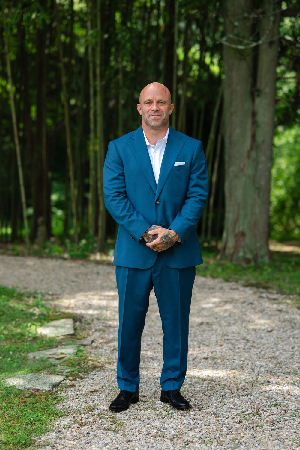 Bald man in a blue suit standing on a gravel path surrounded by lush green trees and bamboo.