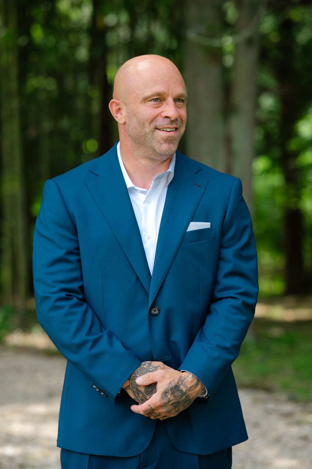 Bald man in blue suit with tattooed hands smiling outdoors among green trees on a sunny day.