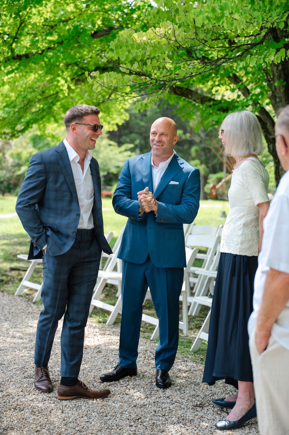 Two men in suits chat outdoors at a wedding ceremony with white chairs and lush green trees.