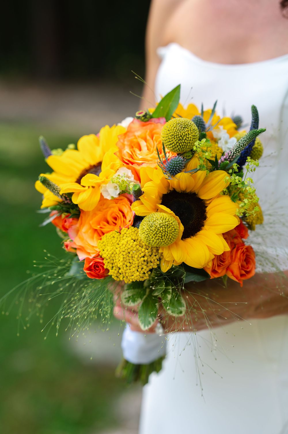 Bride holding vibrant wedding bouquet with sunflowers, orange roses, green craspedia, and blue thistle in yellow and orange tones.