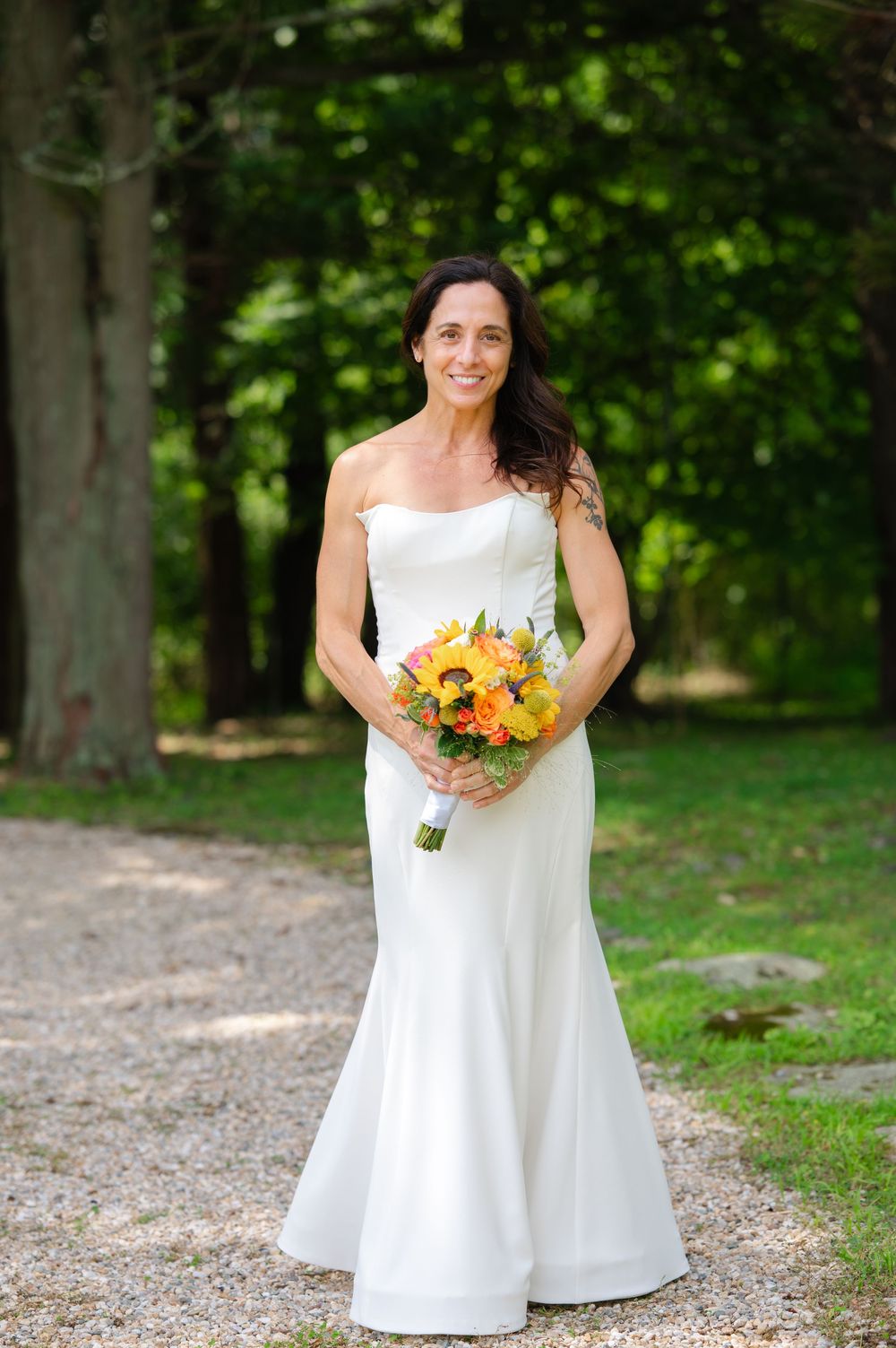 Bride in strapless white mermaid gown holding yellow and orange bouquet, standing on gravel path surrounded by lush green trees.