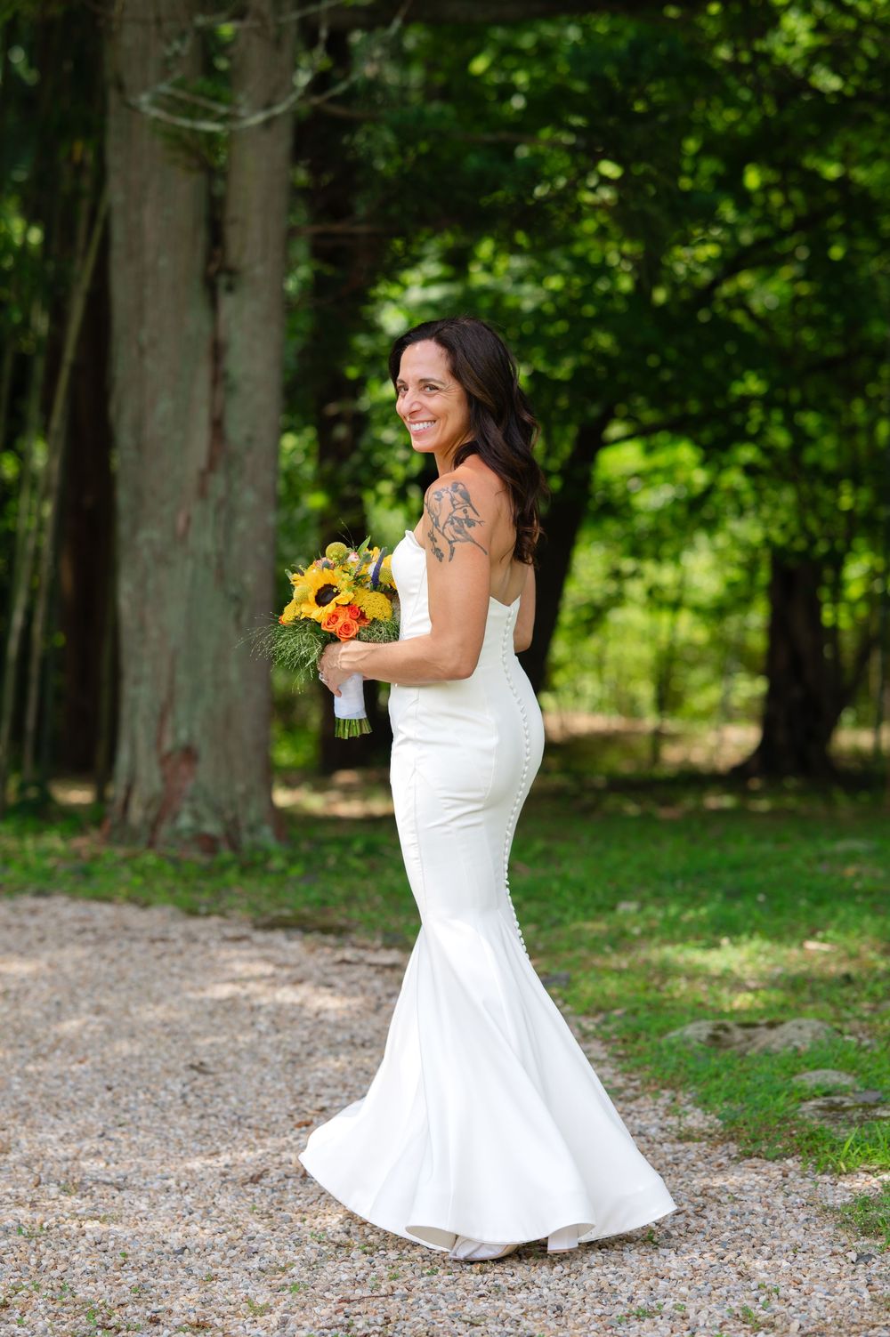 Smiling bride in white mermaid gown holds sunflower bouquet outdoors among lush green trees on gravel path.