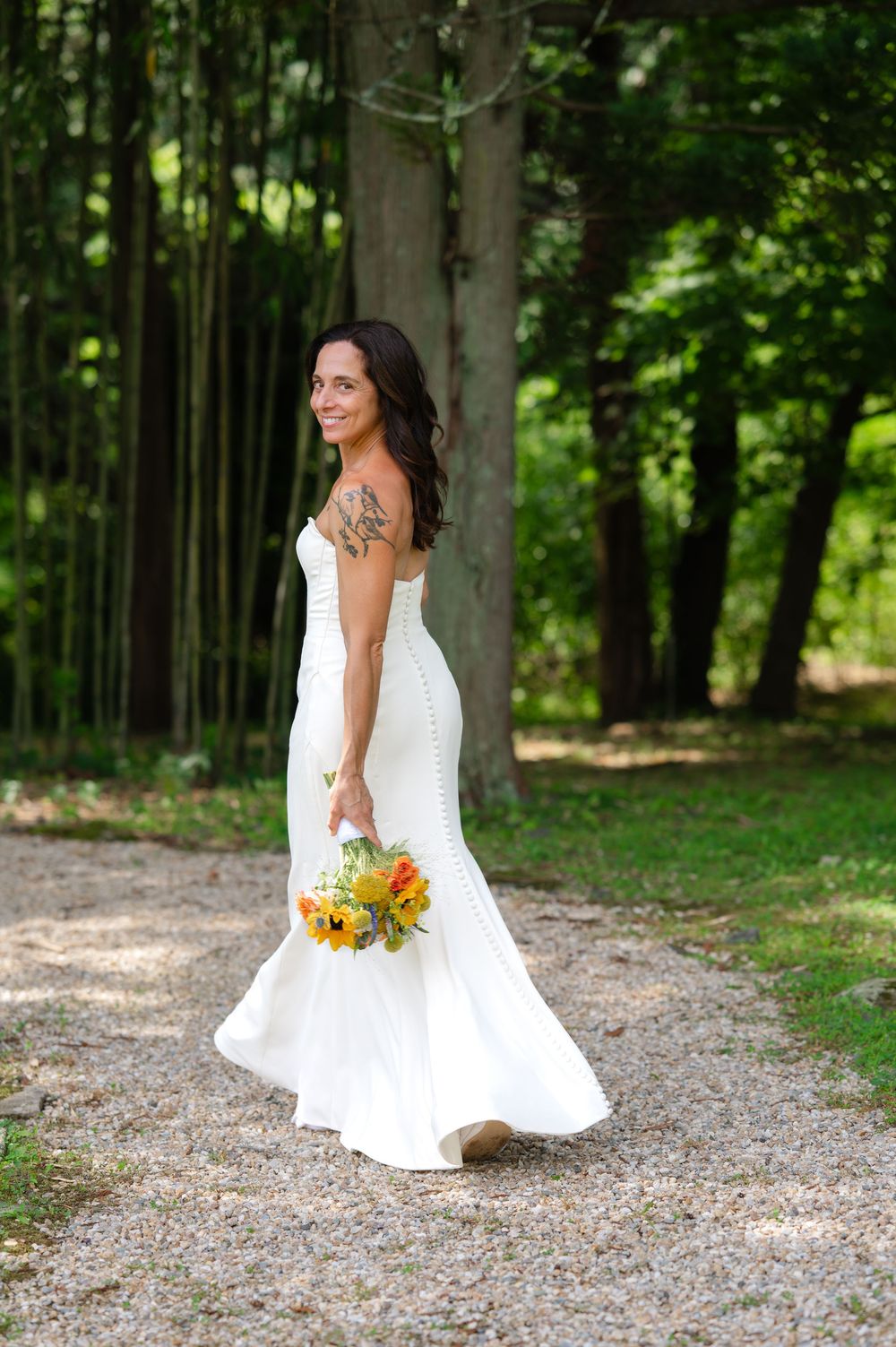 Smiling bride in white mermaid gown holding sunflower bouquet walks along gravel path surrounded by lush green trees.