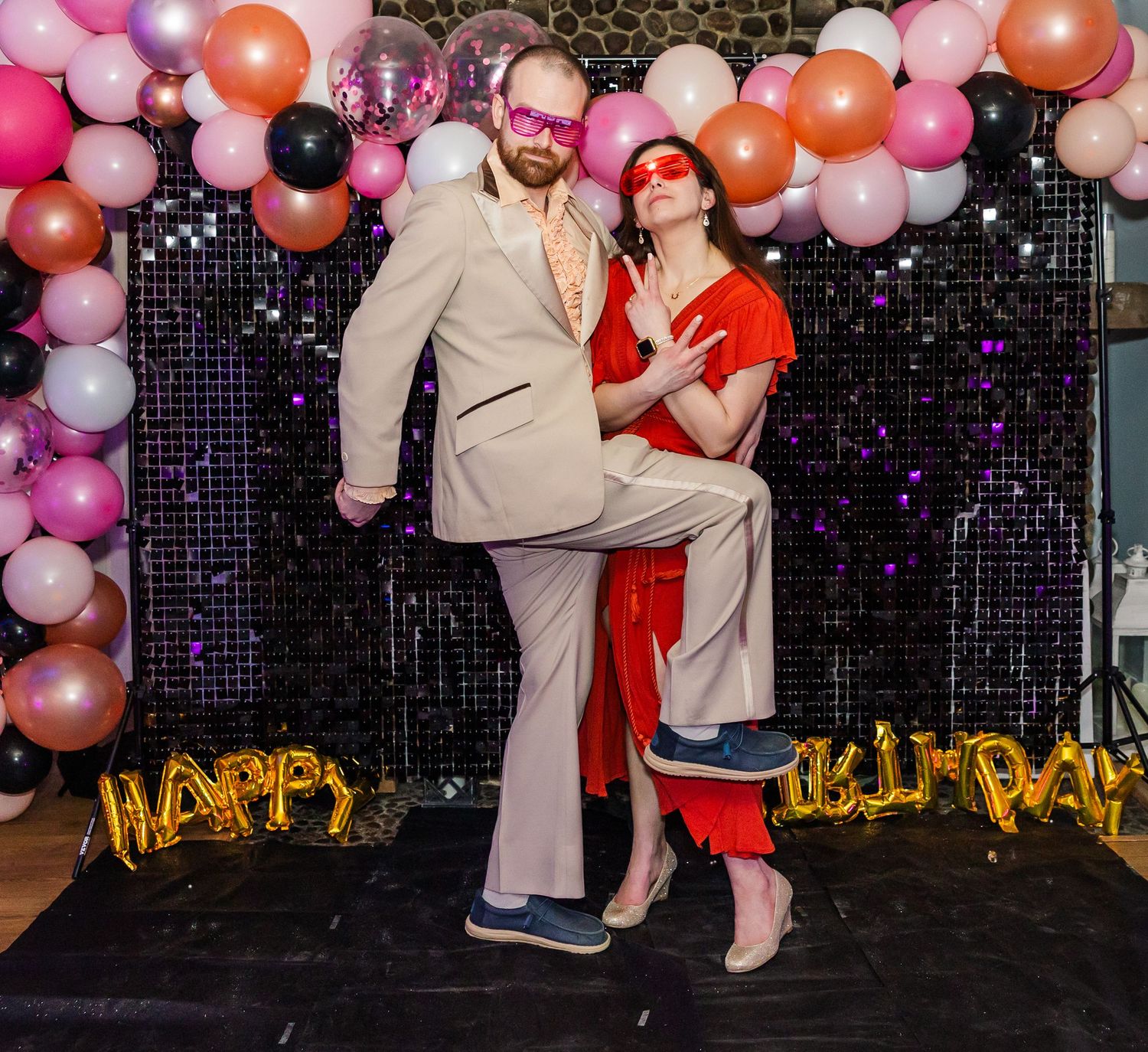 Two people in retro outfits and pink masks pose playfully at a Happy Birthday party with balloon decorations.