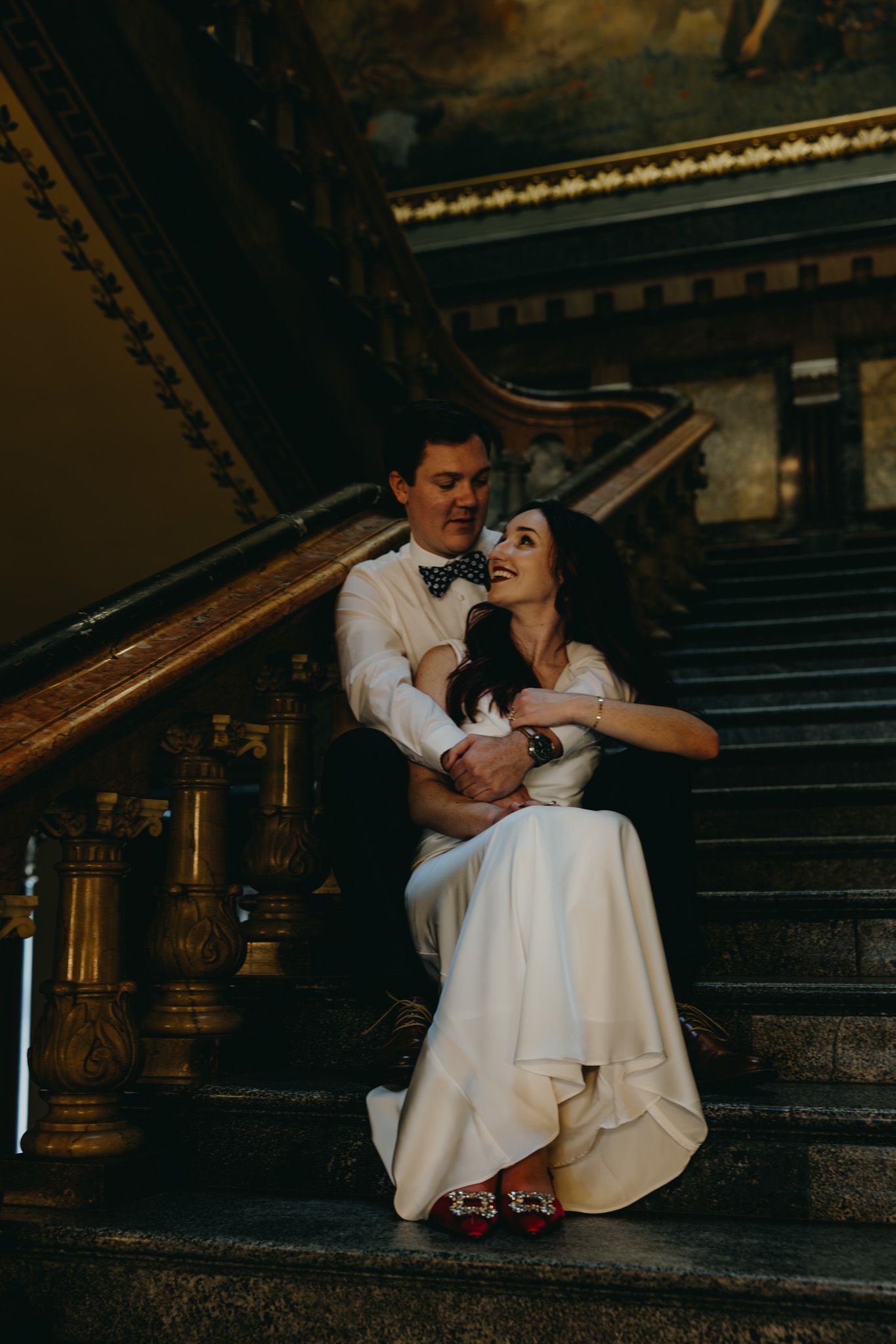 Bride and groom sitting on ornate marble staircase, bride in white gown with red shoes, groom in white shirt and bow tie.