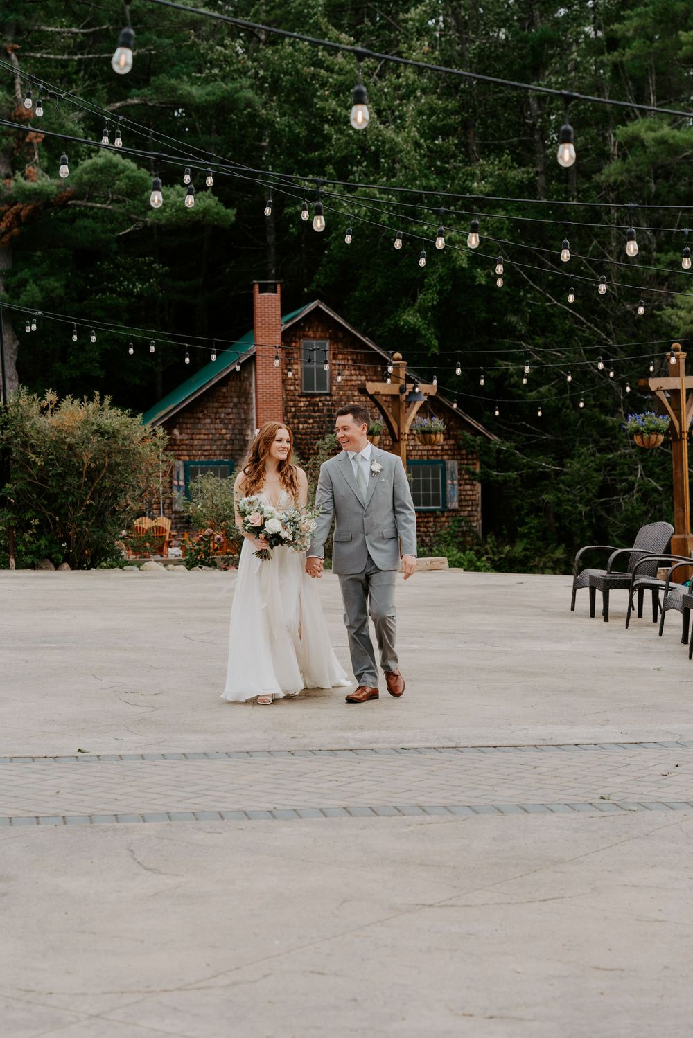 Bride and groom walk hand-in-hand on a stone patio with string lights and a rustic lodge backdrop.