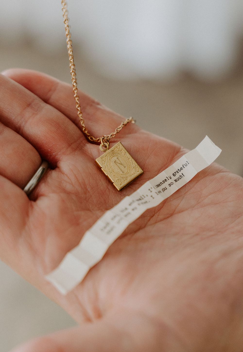Hand holding a gold rectangular locket necklace with a small paper note, on a delicate gold chain.