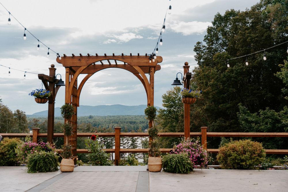 Wooden pergola decorated with string lights and flowers overlooking a scenic mountain and lake landscape at dusk.