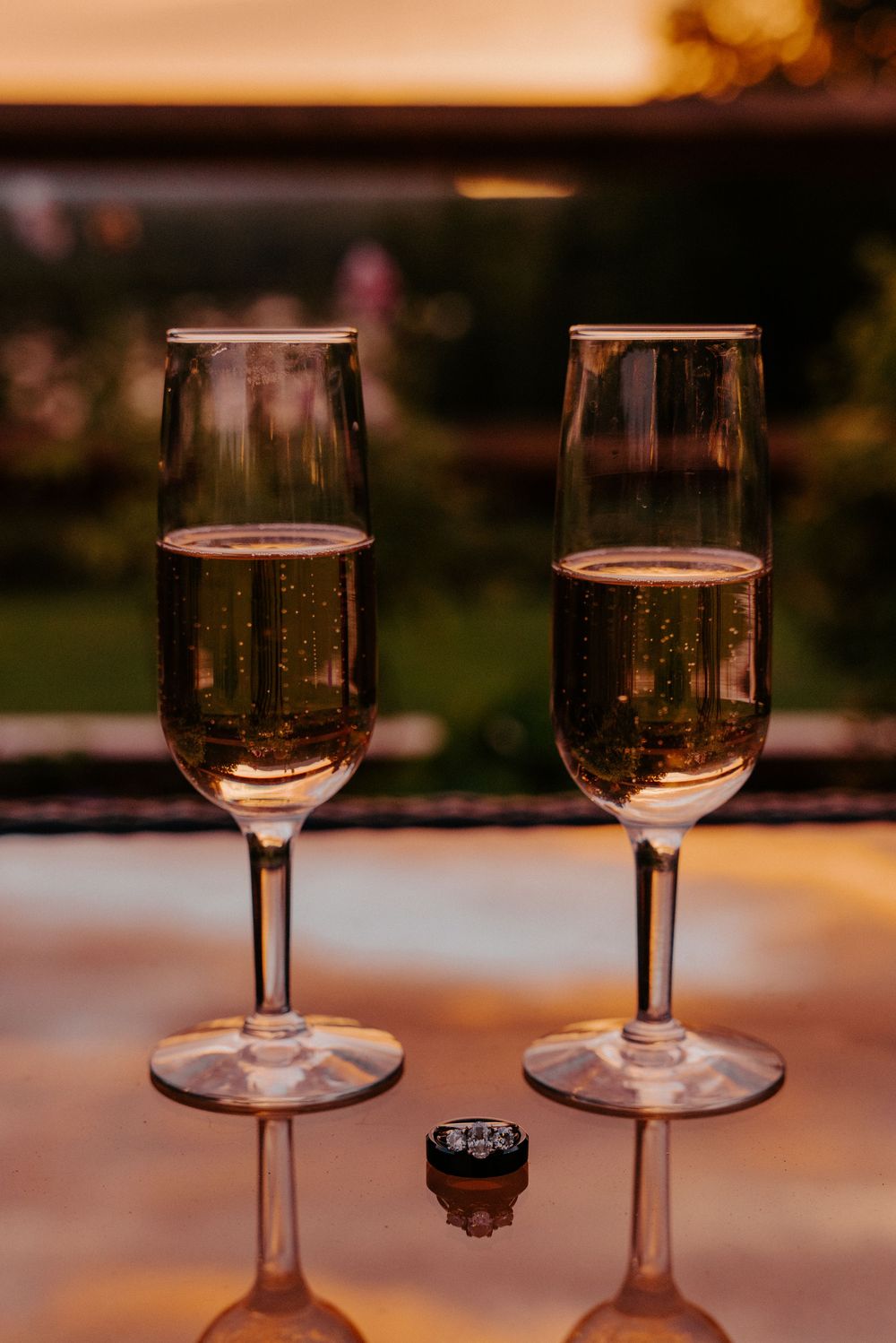Two champagne flutes filled with sparkling wine beside a diamond engagement ring on a reflective surface at sunset.