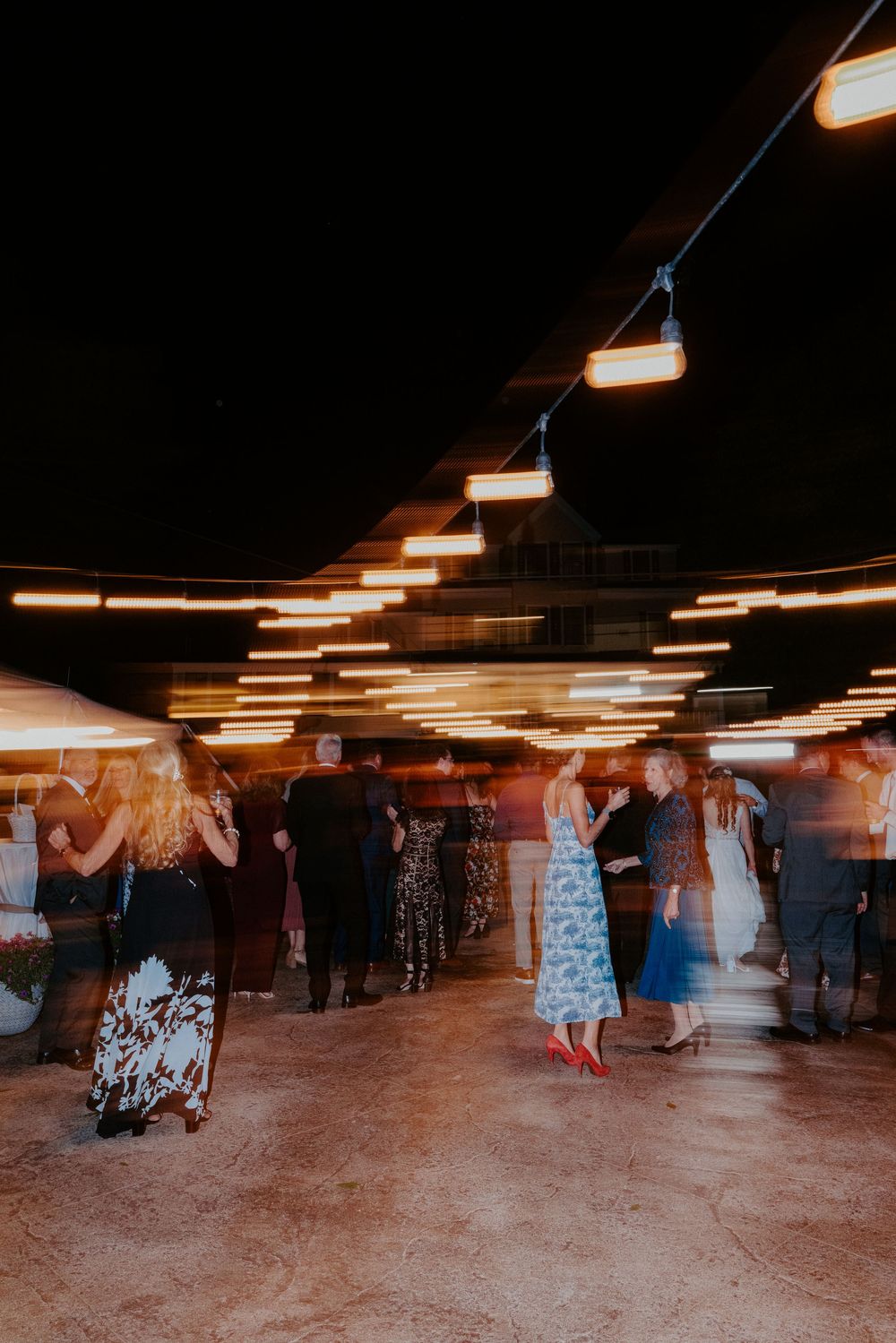 Wedding guests dance under warm string lights in an outdoor tent venue during a lively evening reception.