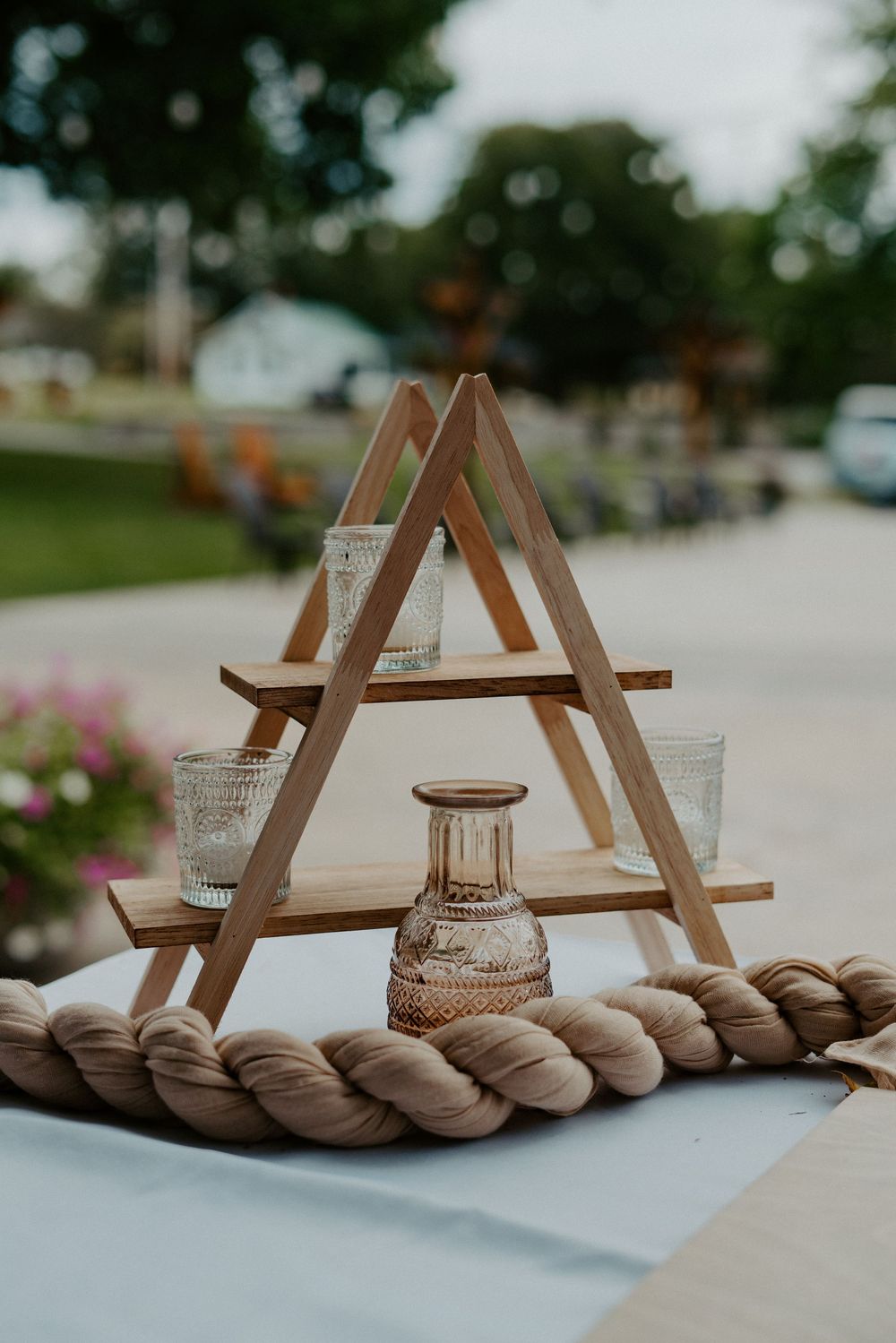 Rustic wooden A-frame tiered display with glass vases and rope detail at outdoor wedding reception.