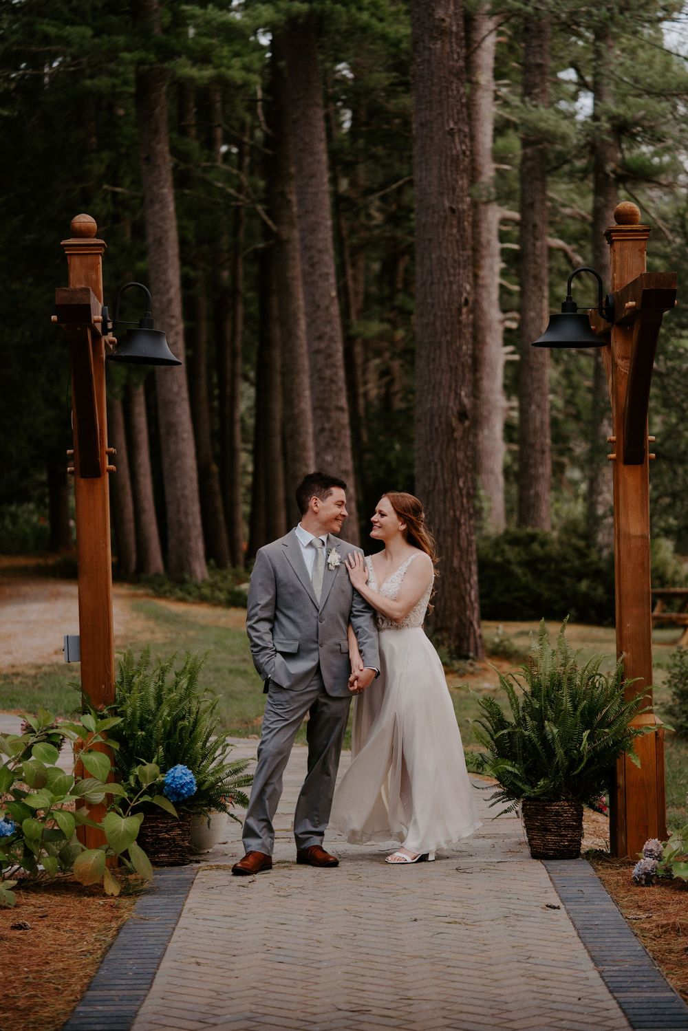 Smiling bride and groom stand together on wooden pathway surrounded by tall pine trees at outdoor wedding venue.