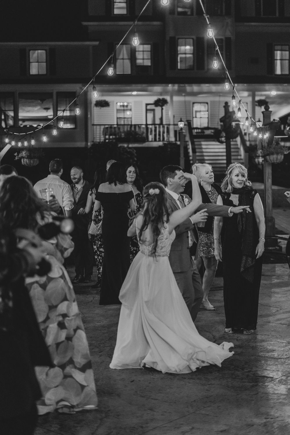 Black and white photo of bride and groom sharing first dance outdoors under string lights with guests.
