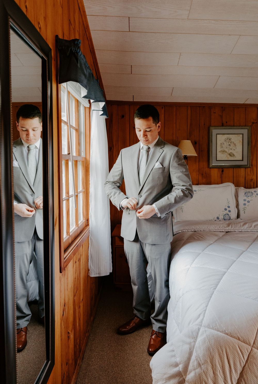 Groom in a light gray suit buttoning his jacket beside a mirror in a cozy wood-paneled bedroom.