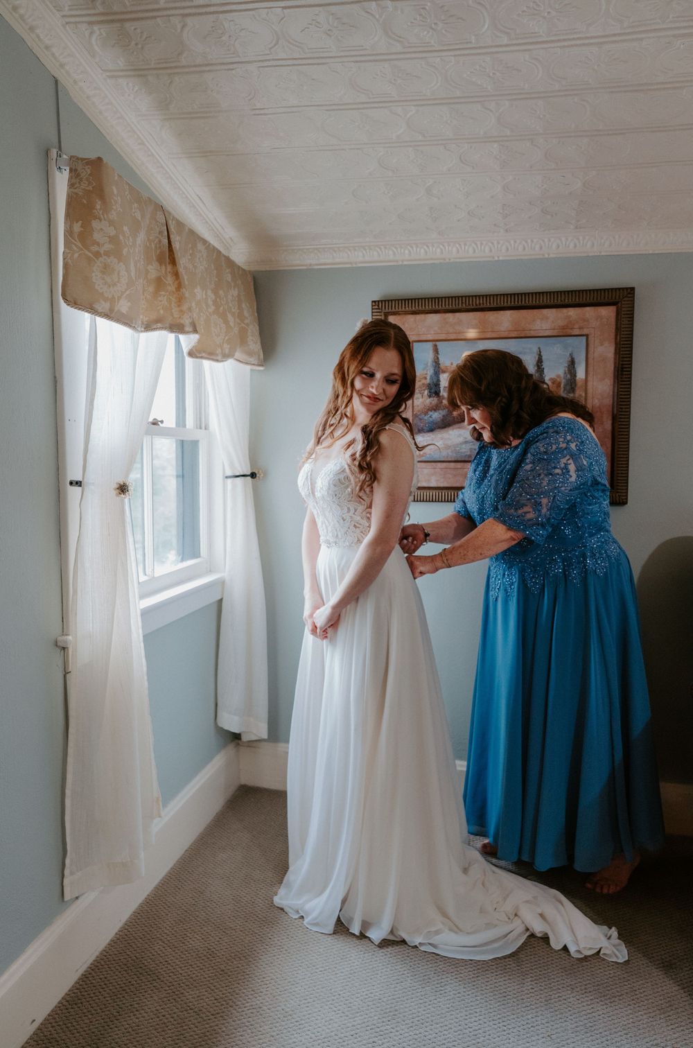 Bride in white gown gets help with dress from woman in blue at window before wedding ceremony.