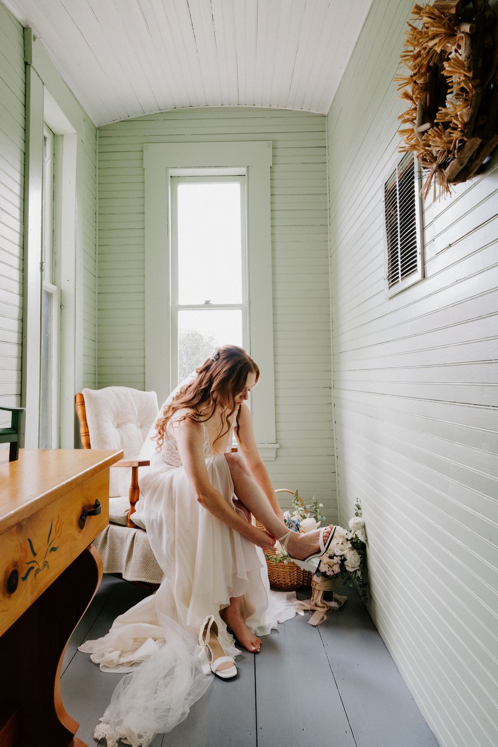 Bride in a flowing white wedding gown putting on her heels in a charming green vintage dressing room.