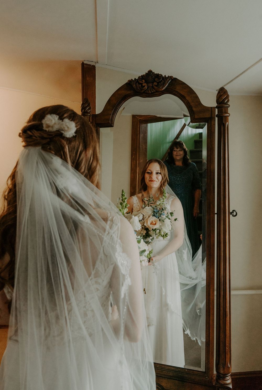 Bride in lace wedding gown and veil admires her reflection in an ornate wooden floor mirror while holding a bouquet.