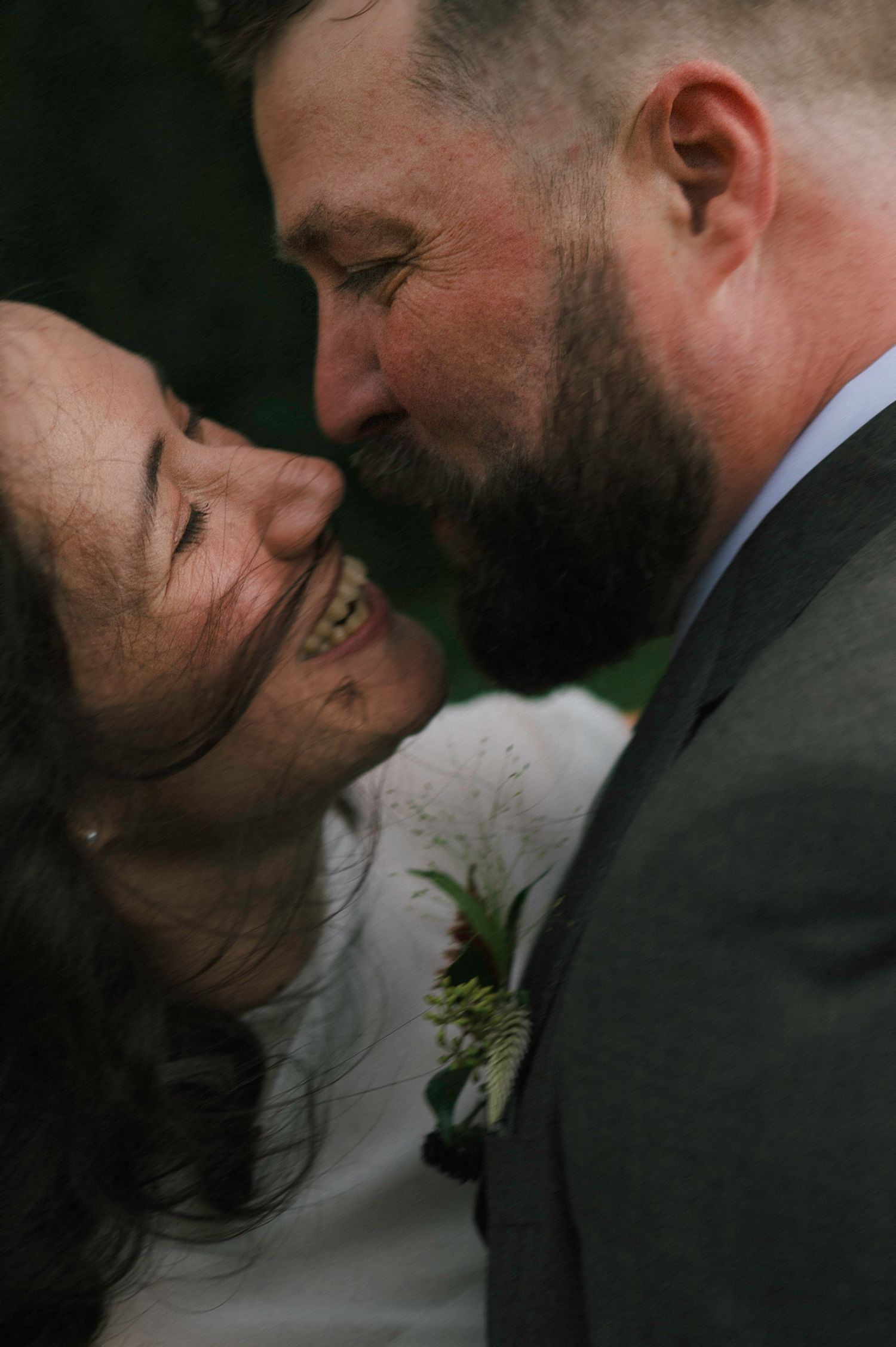 A smiling bride and bearded groom share a tender moment, nearly kissing, with the groom wearing a boutonniere on his suit.