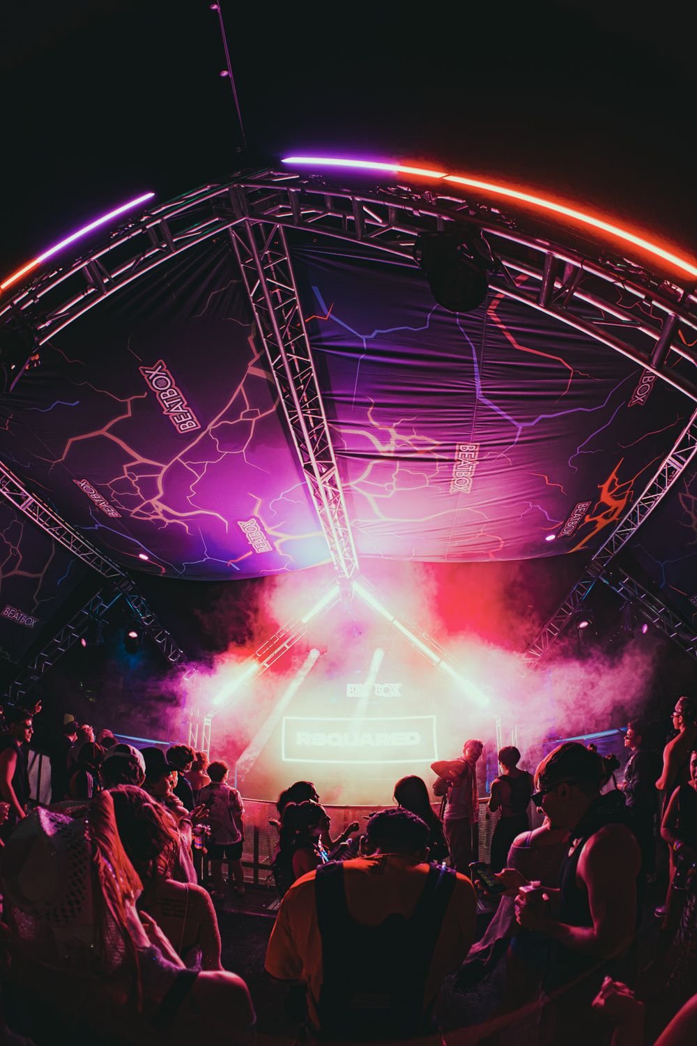 Crowd dancing under vibrant neon lights at an outdoor music festival stage with pink and purple lighting.