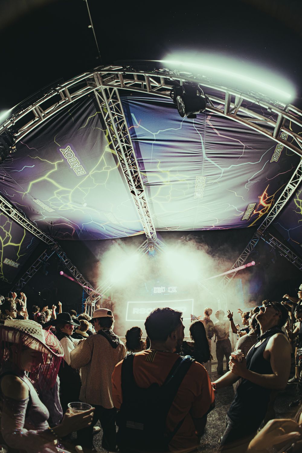 Crowd of festival-goers dance under a large illuminated stage with fog and lightning bolt visuals at an outdoor music event.
