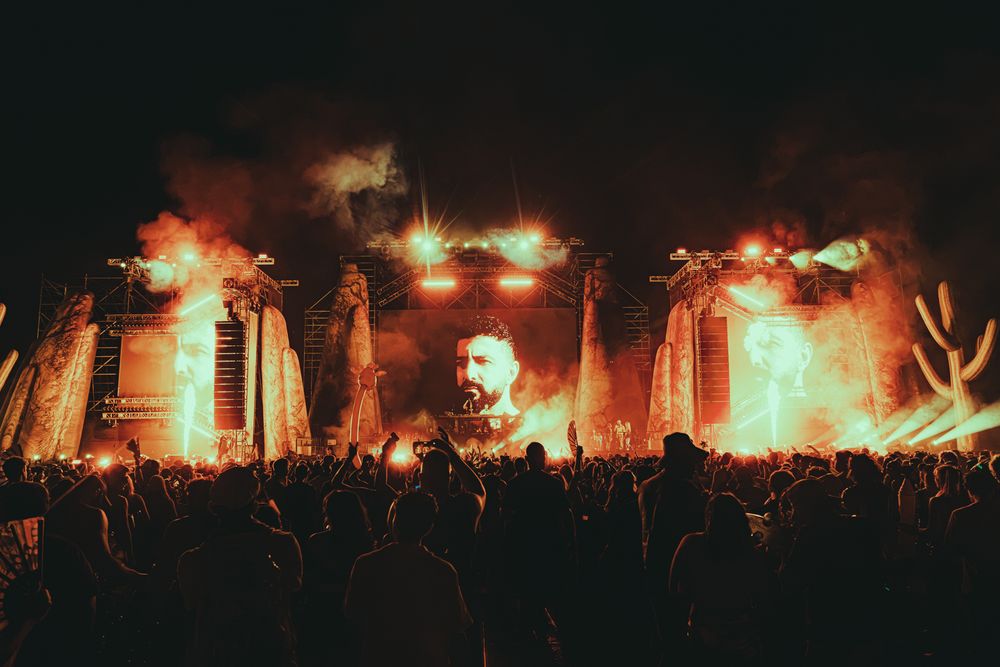 Spectacular outdoor concert stage with fiery red and orange pyrotechnics lighting up a massive nighttime crowd.