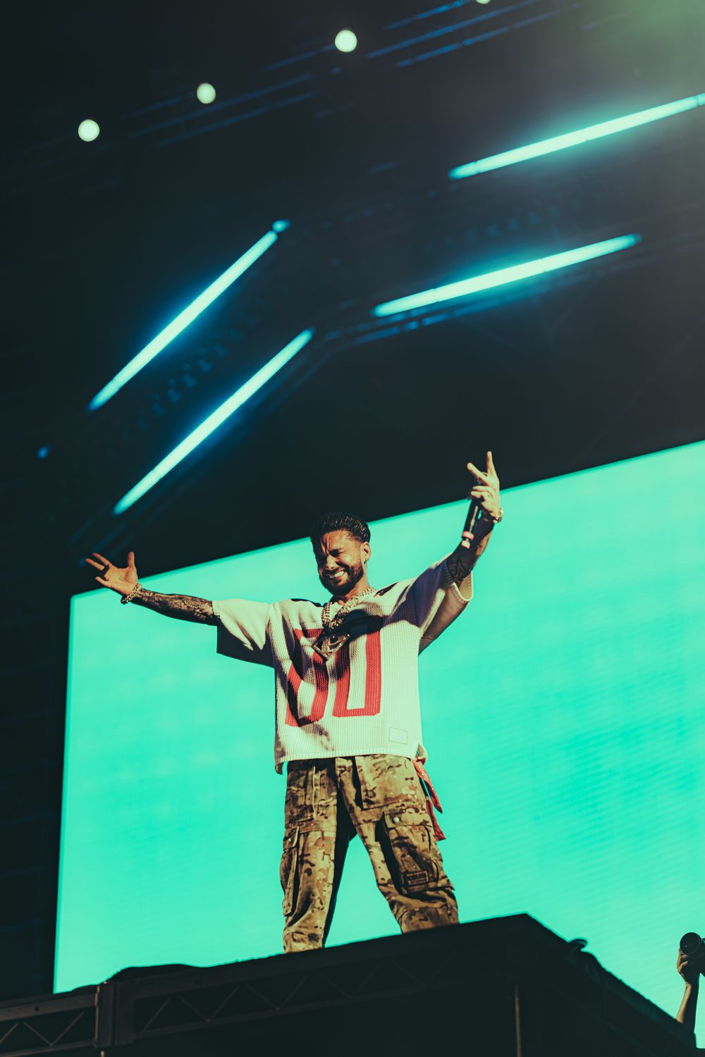 Performer on stage wearing jersey and cargo pants, raising hands with peace sign before green LED backdrop.