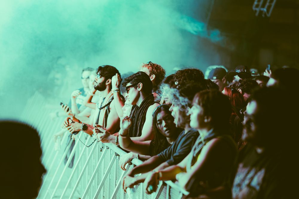 Concert crowd standing behind barriers under green atmospheric stage fog at a live music event.