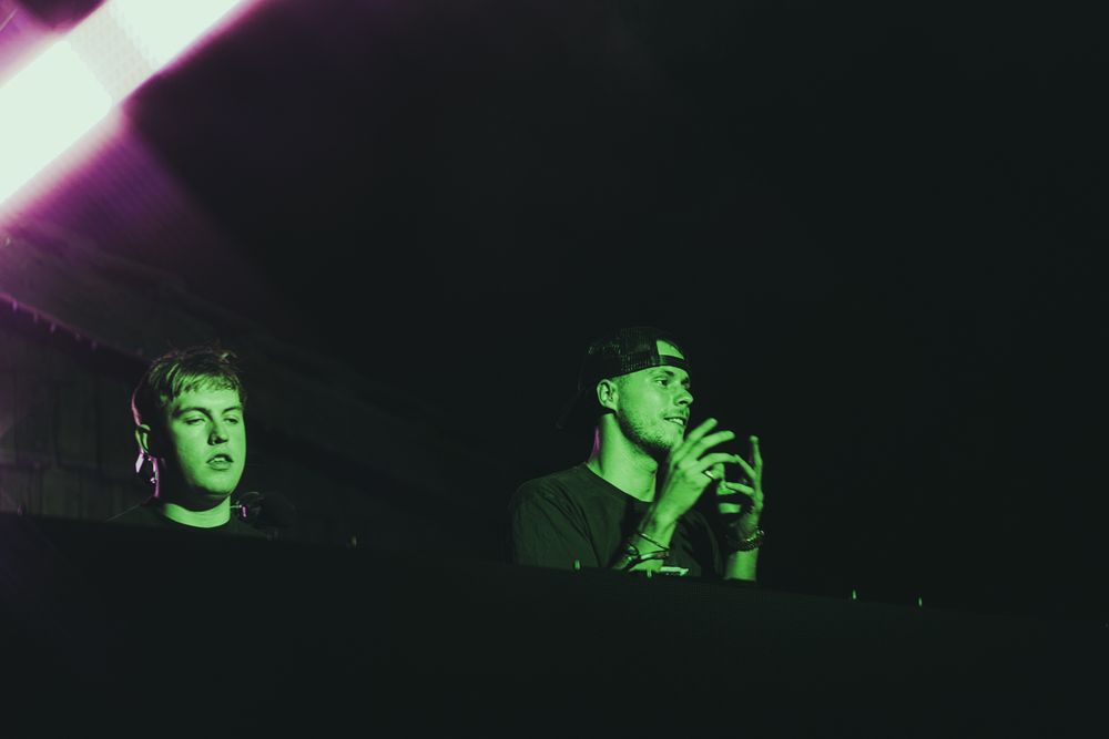 Two male DJs behind a booth bathed in green light in a dark performance venue.