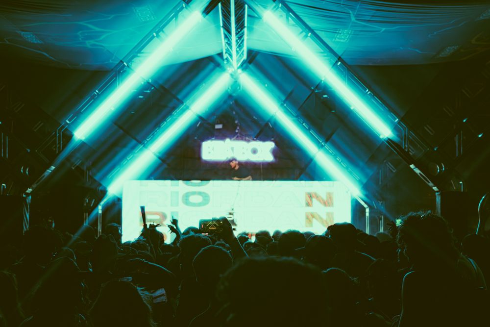 Crowd at an indoor electronic music festival with neon green geometric lights and a DJ stage in the background.