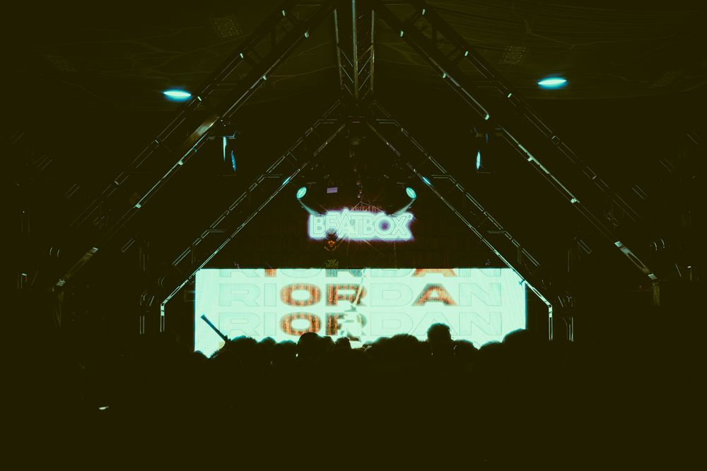 Dark concert venue with illuminated screen displaying text, silhouetted crowd, and dramatic stage lighting structure overhead.