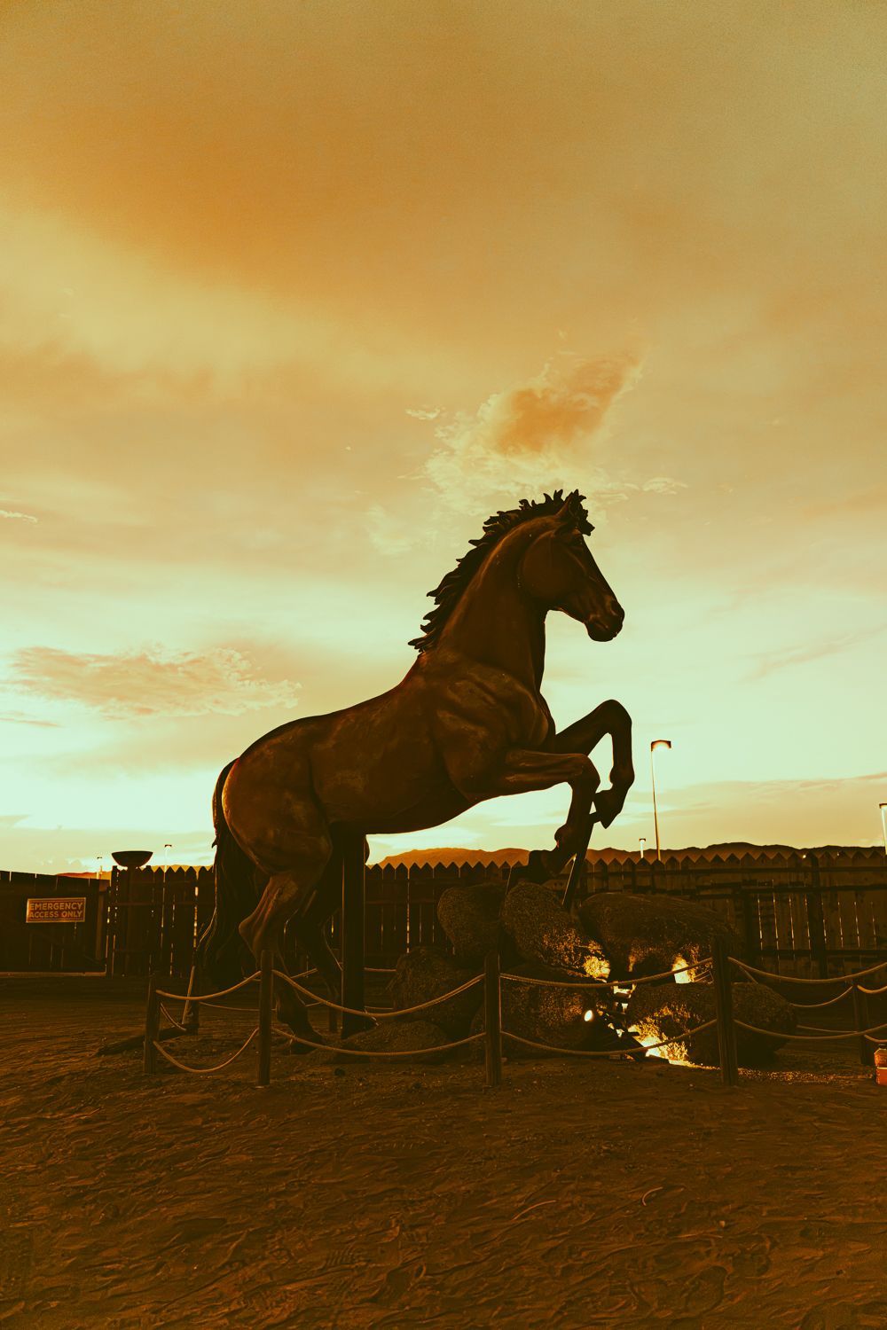Dramatic silhouette of a rearing horse statue against a golden sunset sky in an outdoor arena setting.