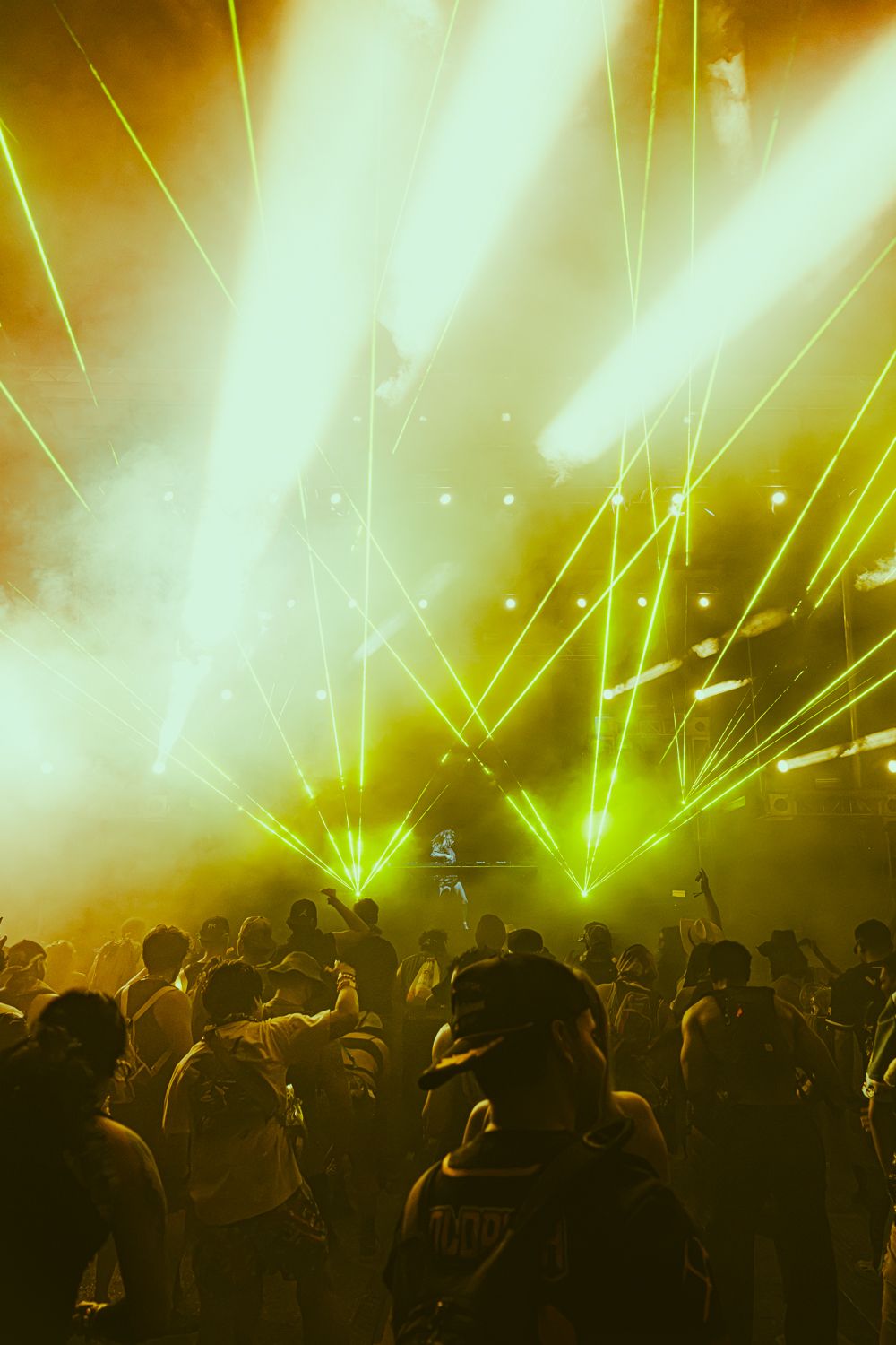 Dense crowd at a nightclub event illuminated by intense yellow and green laser light beams through smoke.