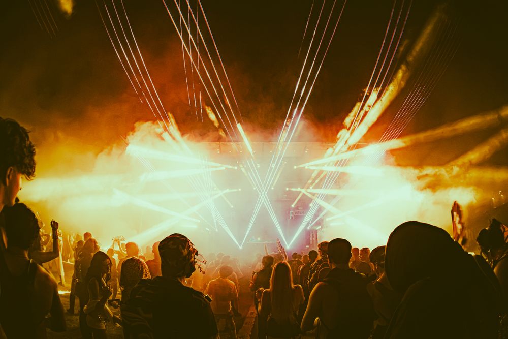 Crowd silhouettes at an outdoor music festival with dramatic laser lights and smoke effects illuminating the night sky.