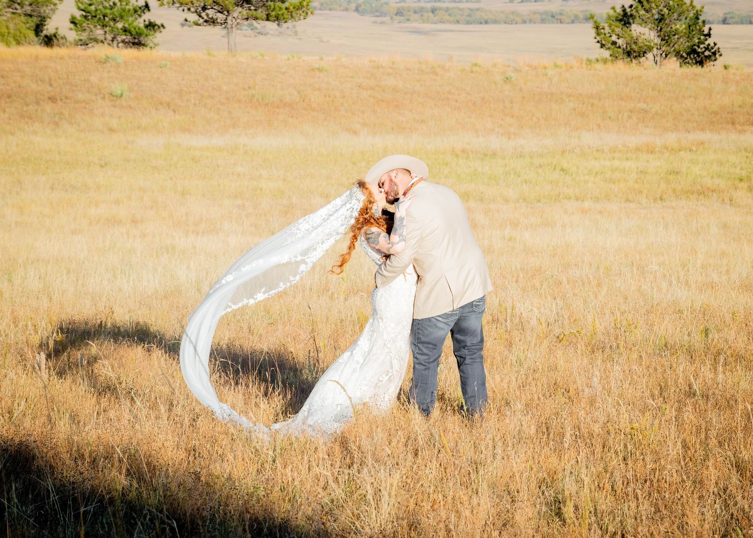 Bride and groom share a romantic kiss in a golden field, her veil flowing in the breeze on their wedding day.