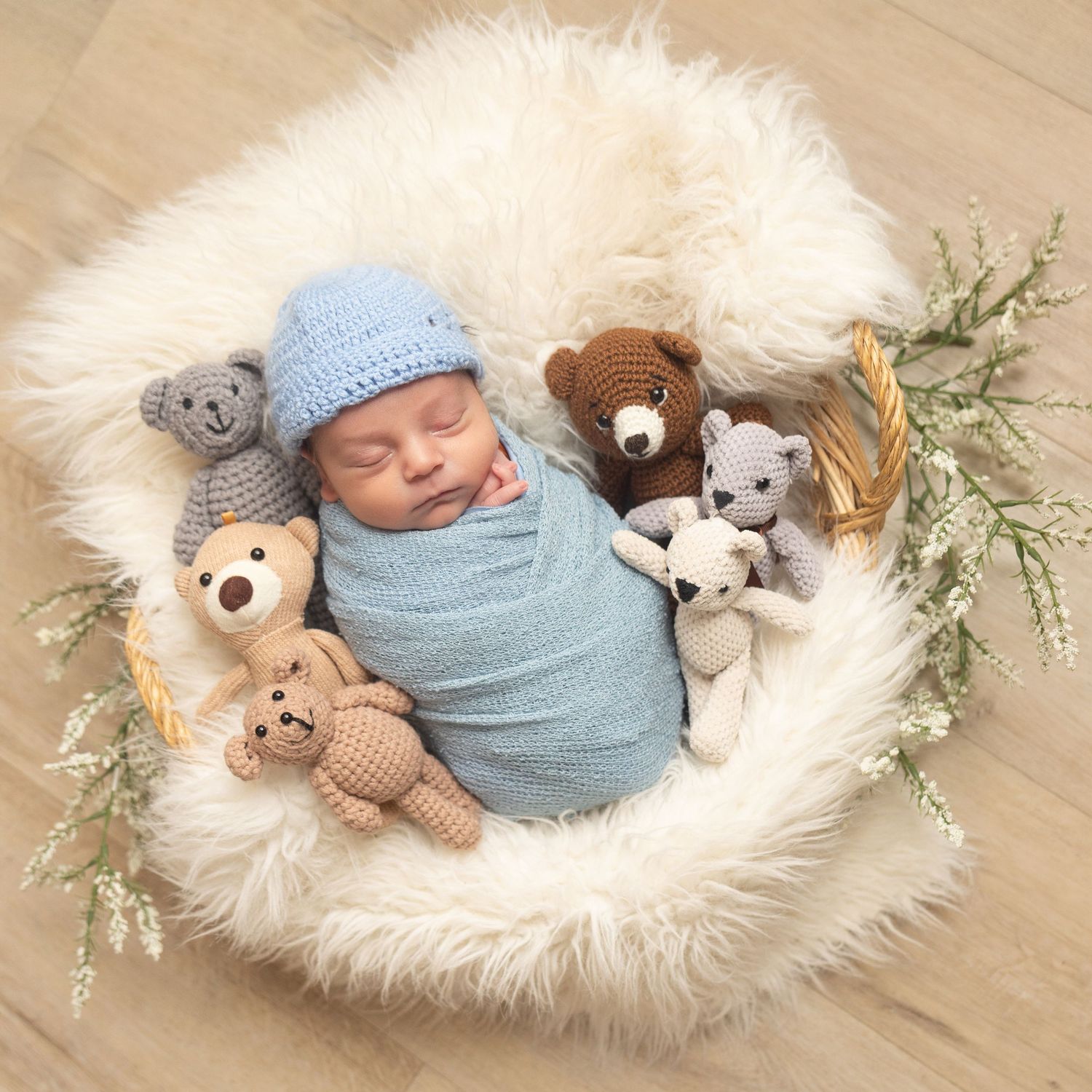 Sleeping newborn baby wrapped in blue, wearing a knit hat, surrounded by crocheted teddy bears in a fluffy white basket.