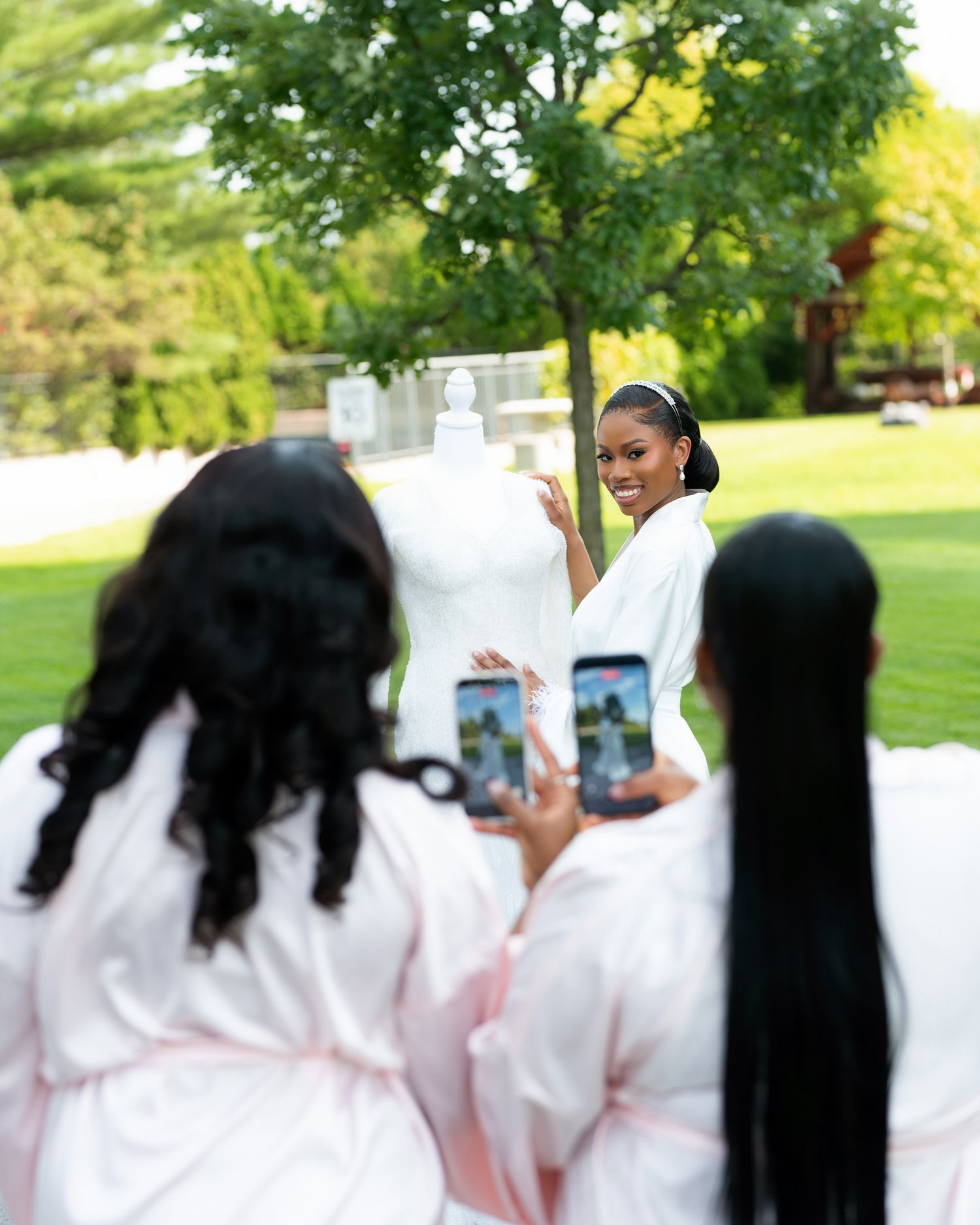Bridesmaids in white robes photograph a smiling bride posing beside her wedding dress on a lush green lawn.