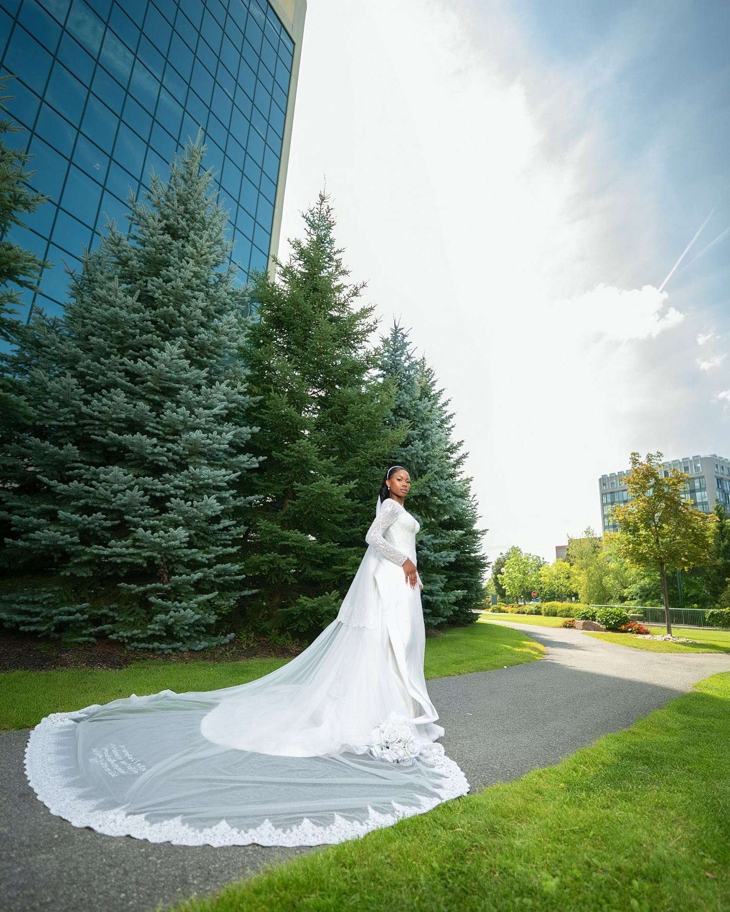 Bride in elegant long-sleeve white gown with cathedral veil on garden path beside tall evergreens and modern glass building.