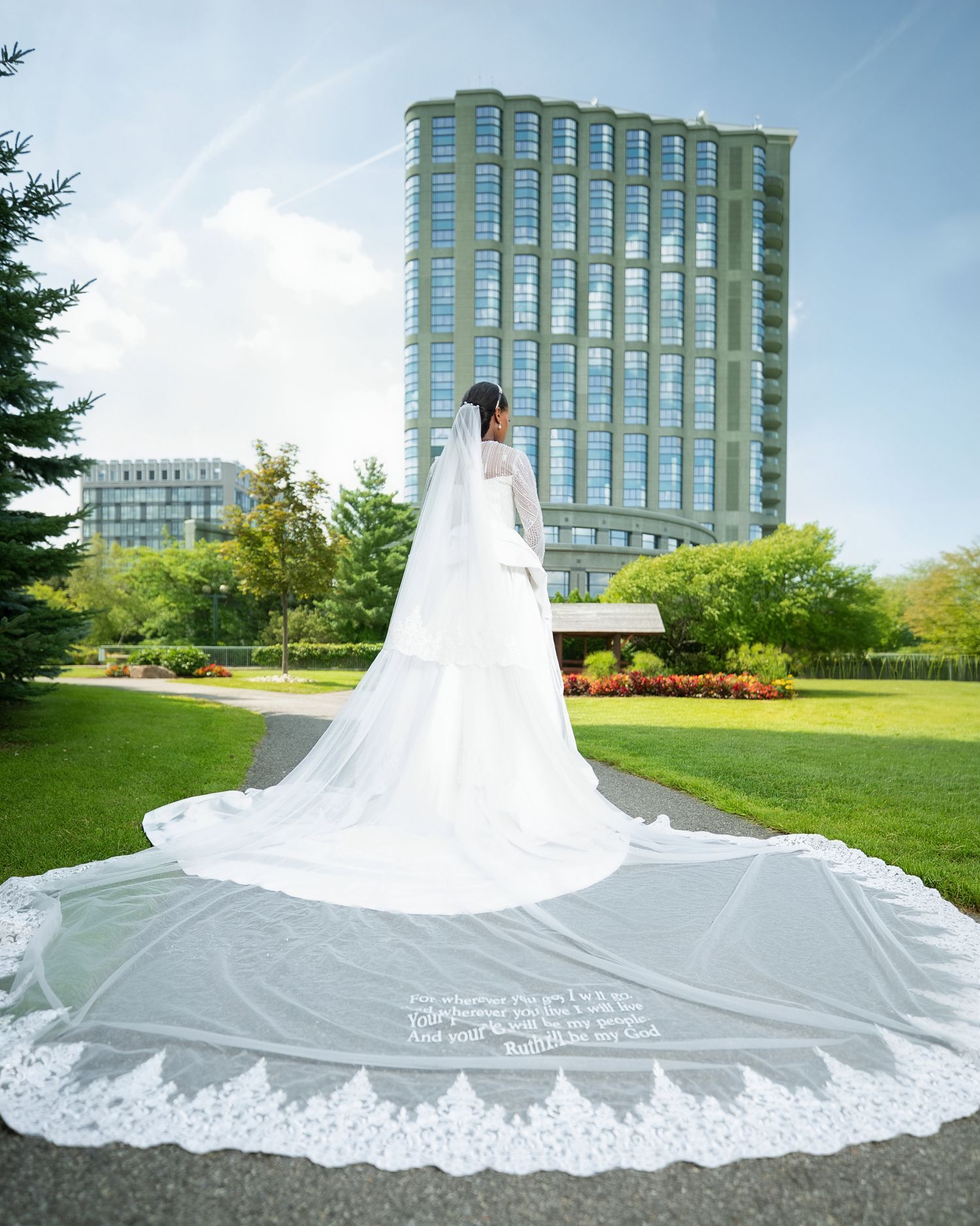 Bride in white gown with long lace-trimmed cathedral veil stands in park, modern glass building backdrop.