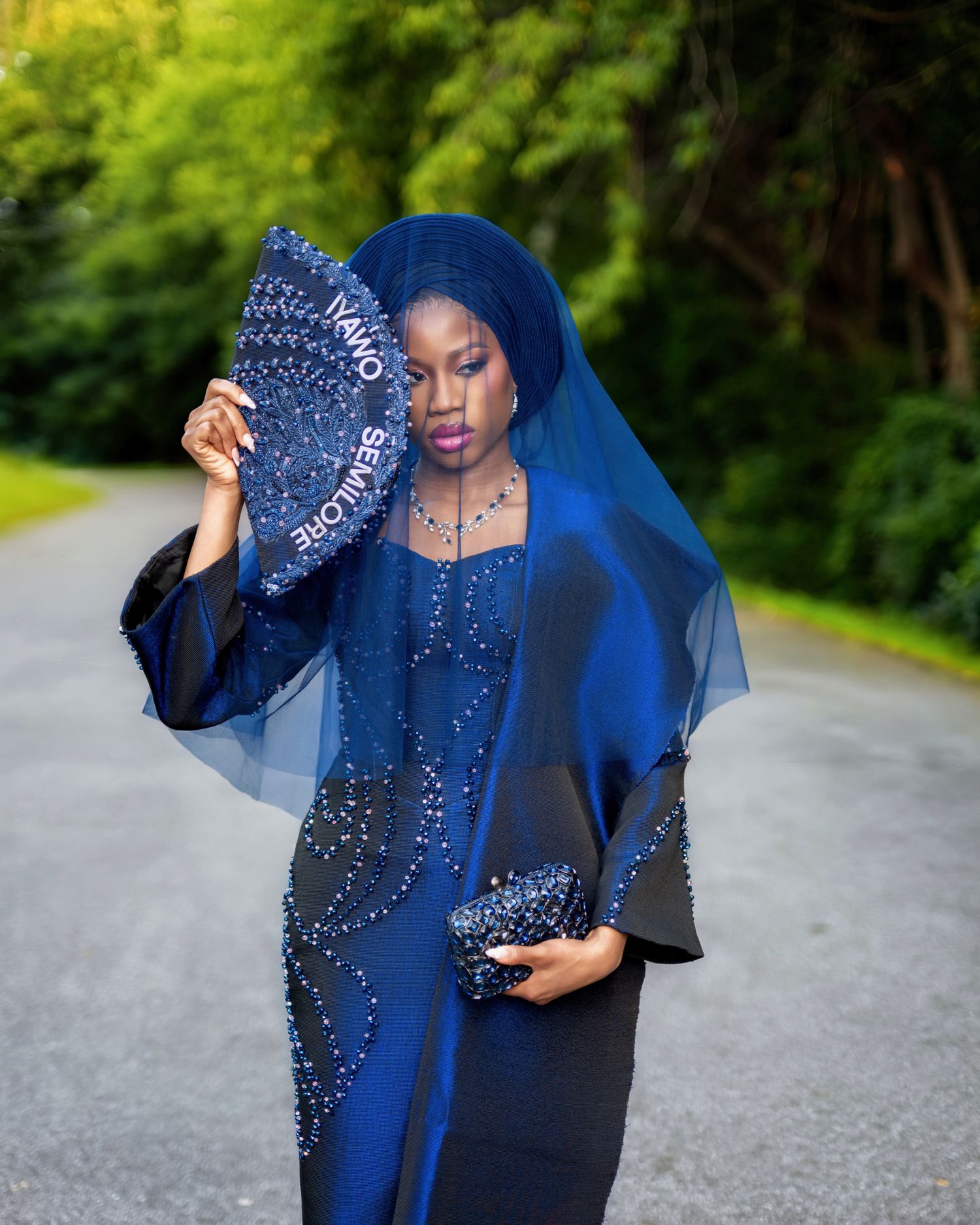 Elegant woman in royal blue beaded gown with sheer veil holding decorative fan outdoors.