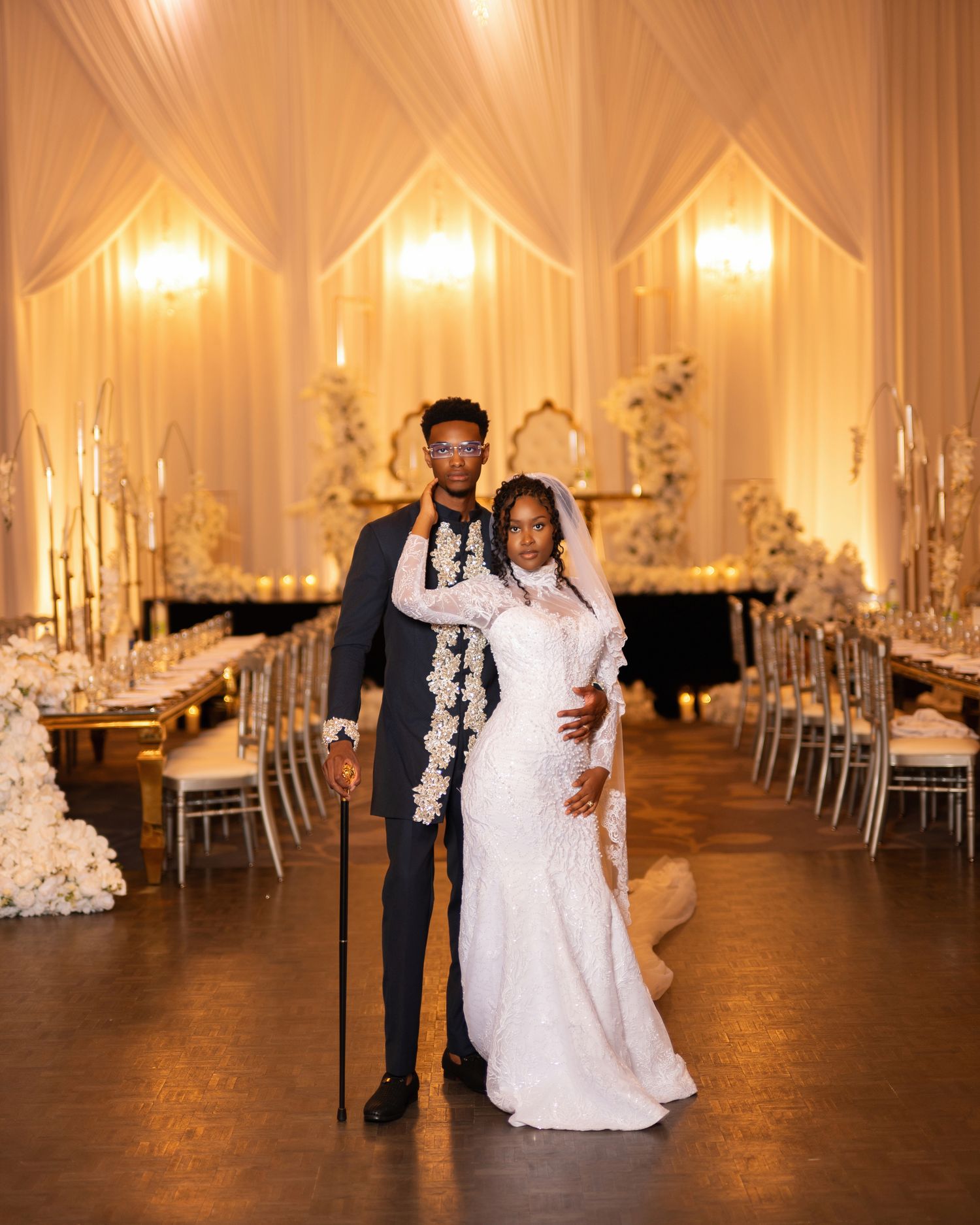 Elegant Black couple poses in a grand wedding reception hall with white floral decor, draped ceilings, and golden candlelight.