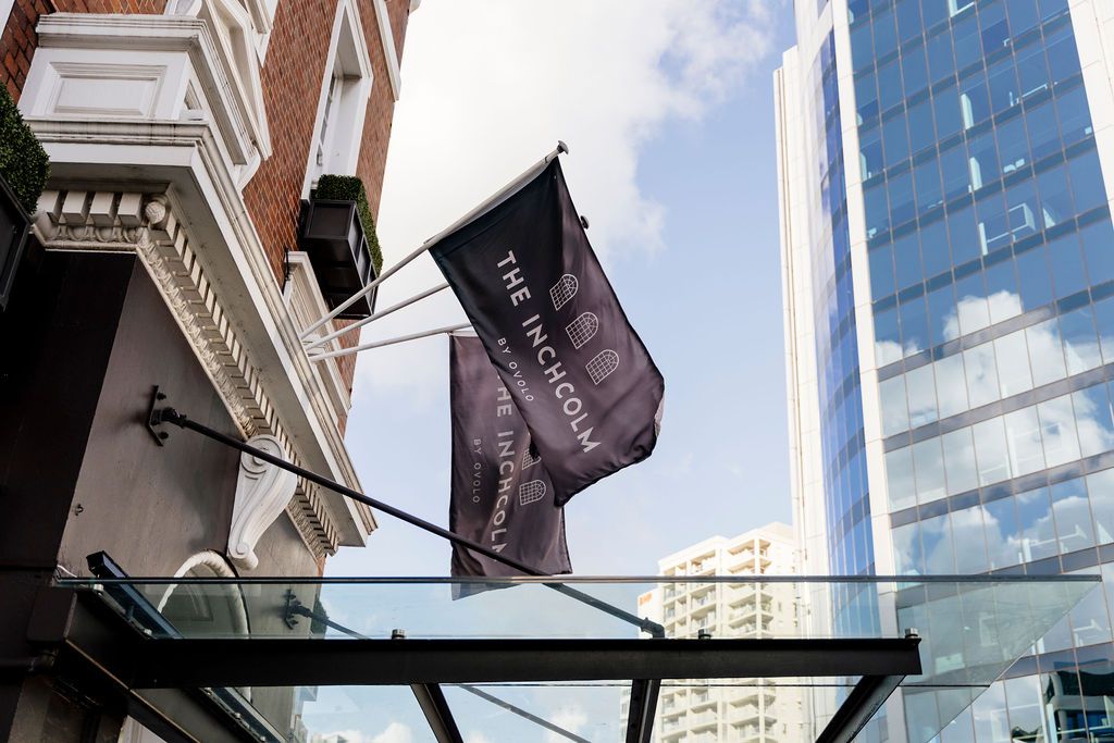 The Inchcolm hotel entrance with branded flags on a glass canopy between historic brick and modern glass buildings.