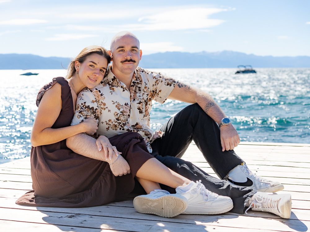 Couple sitting together on a wooden dock by a sparkling lake with mountains in the background.