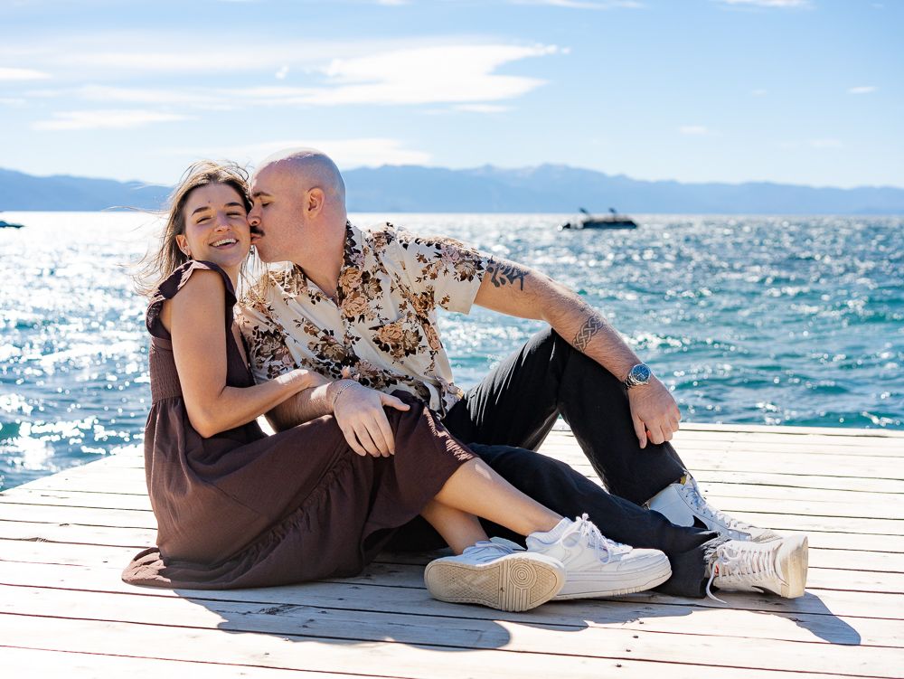 Couple sitting on a sunny lakeside dock, man kissing woman's cheek, mountains in background.