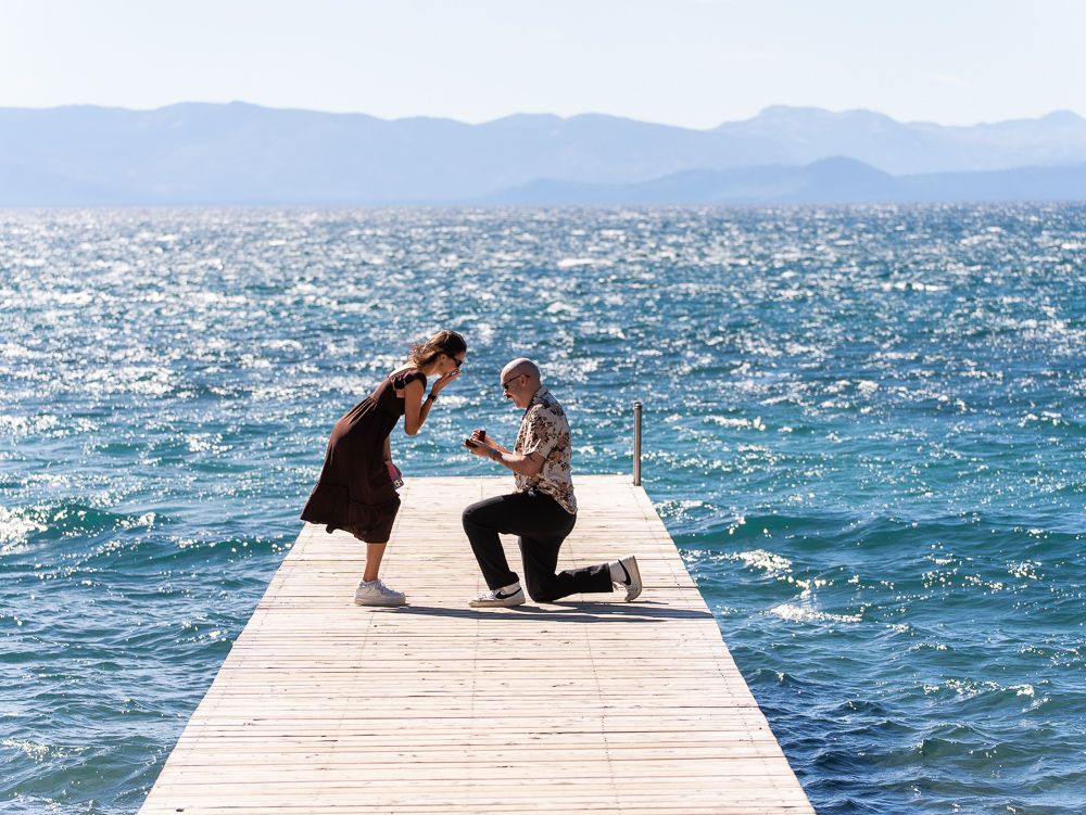 Man proposing on a wooden dock over sparkling blue lake with mountain backdrop on a sunny day.
