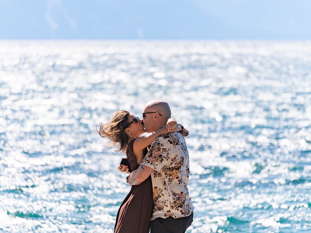 Couple sharing a romantic kiss by the sparkling blue ocean, with mountains visible in the background.