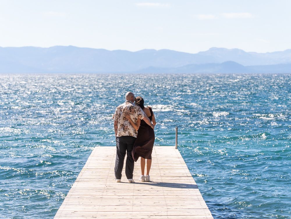 Couple walking arm in arm on a wooden pier overlooking a sparkling blue lake with mountain views.