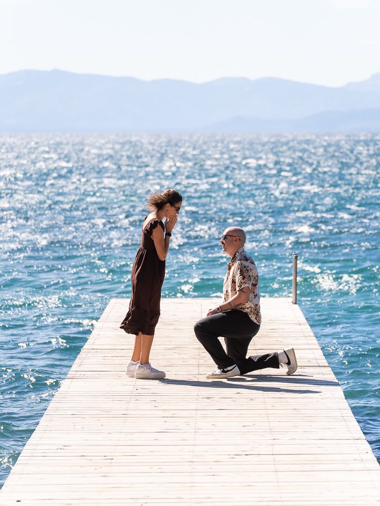 Person proposing on a wooden dock over sparkling blue lake water with mountains in the background.