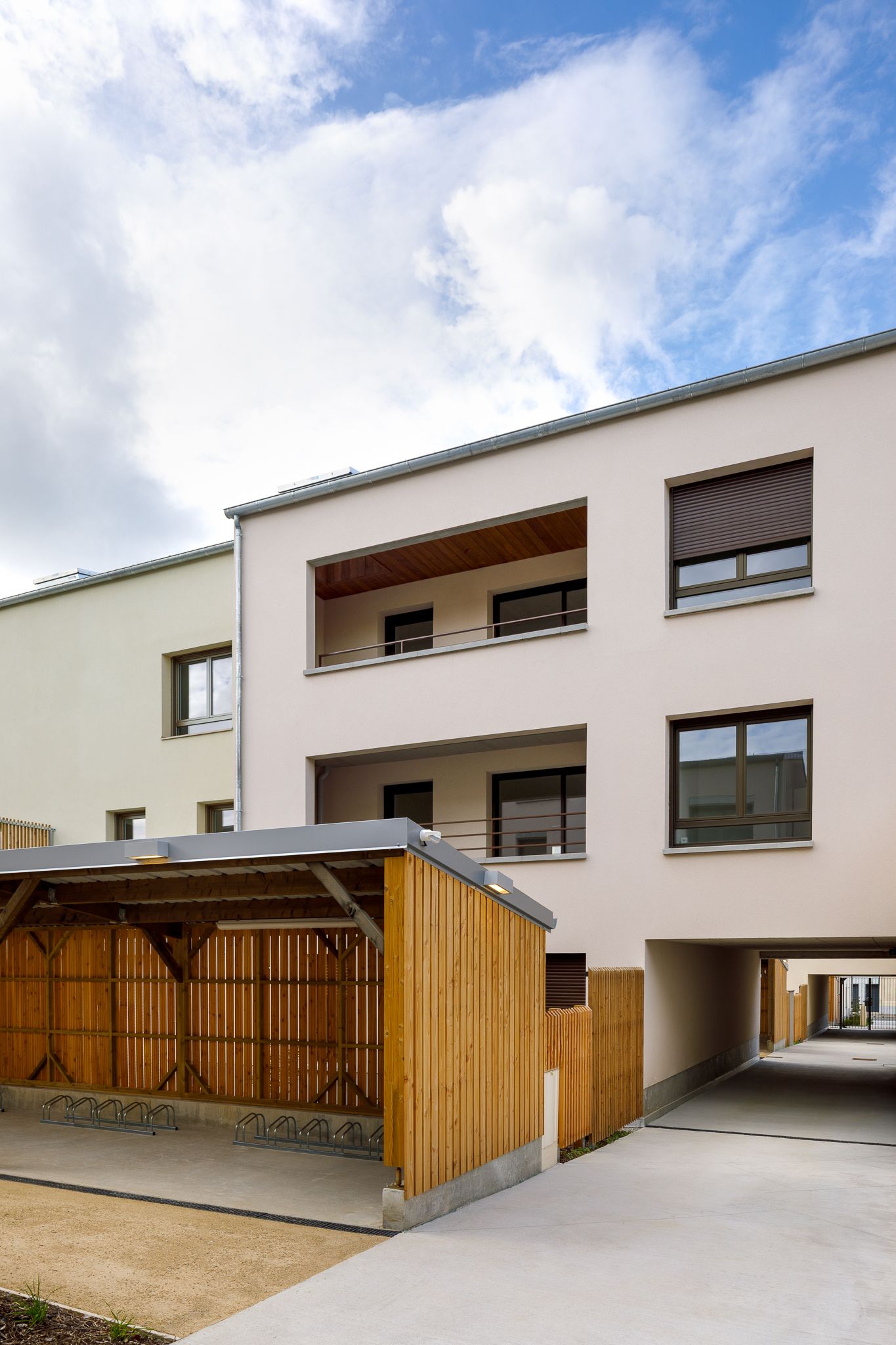 Immeuble résidentiel de couleur beige, doté d'un abri vélo en bois, de balcons et d'une entrée de parking souterrain.