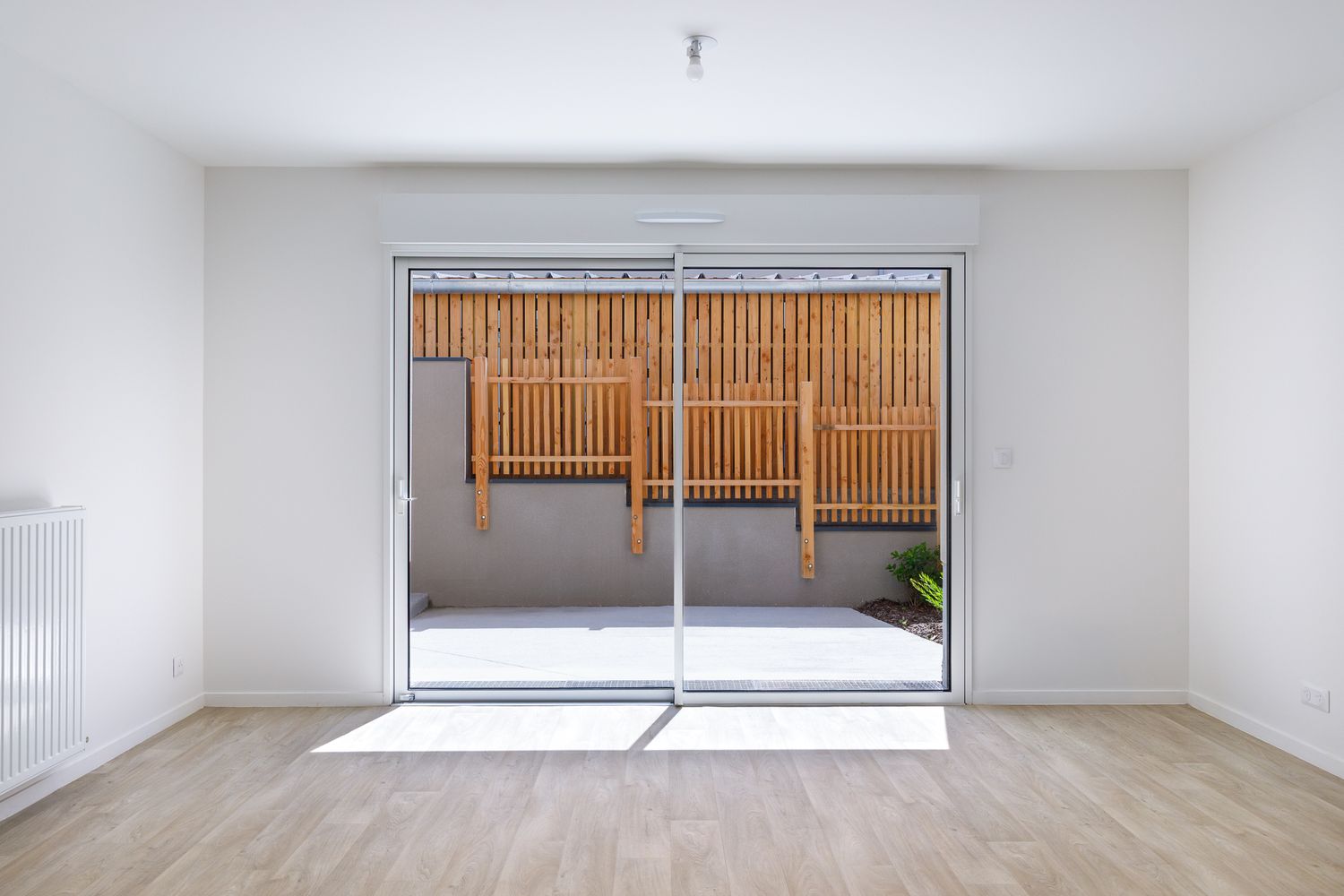 Pièce lumineuse et vide, dotée de portes-fenêtres coulissantes s'ouvrant sur un balcon en bois, avec des sols clairs.